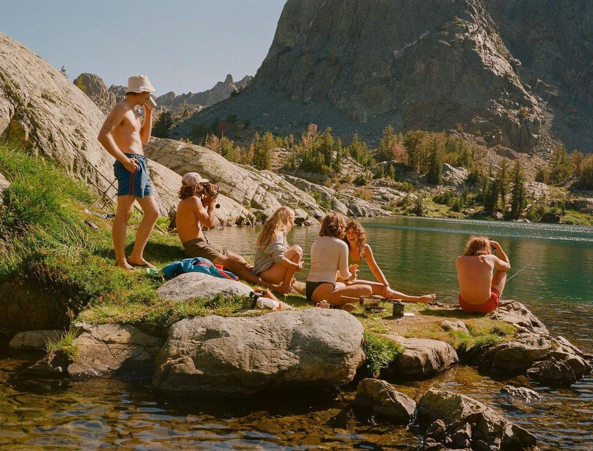 Six friends by a mountain lake, one pouring coffee, one filming